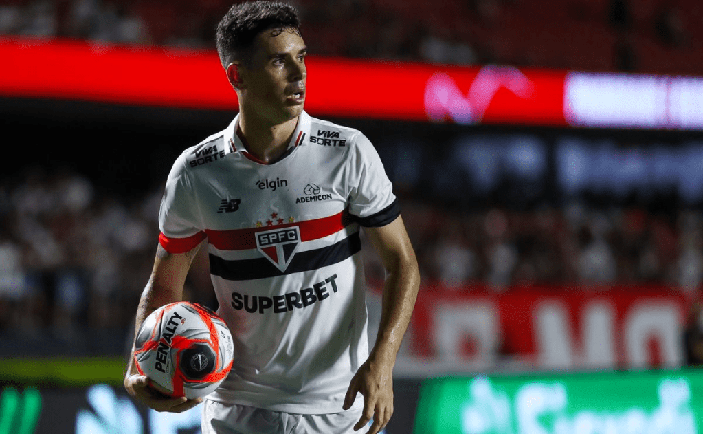 Jogador de futebol do São Paulo FC segurando uma bola em campo, vestido com uniforme do time, em um estádio lotado.