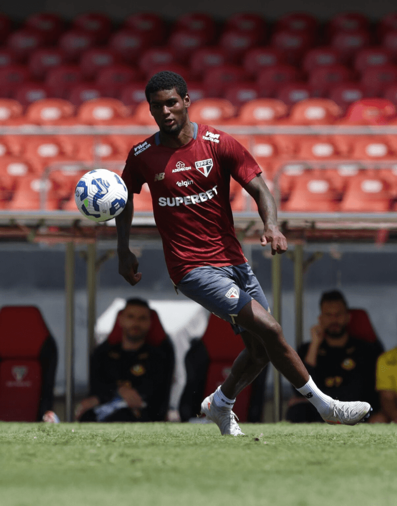 Jogador de futebol em um treino, correndo em campo e segurando a bola, com bancadas vermelhas ao fundo.