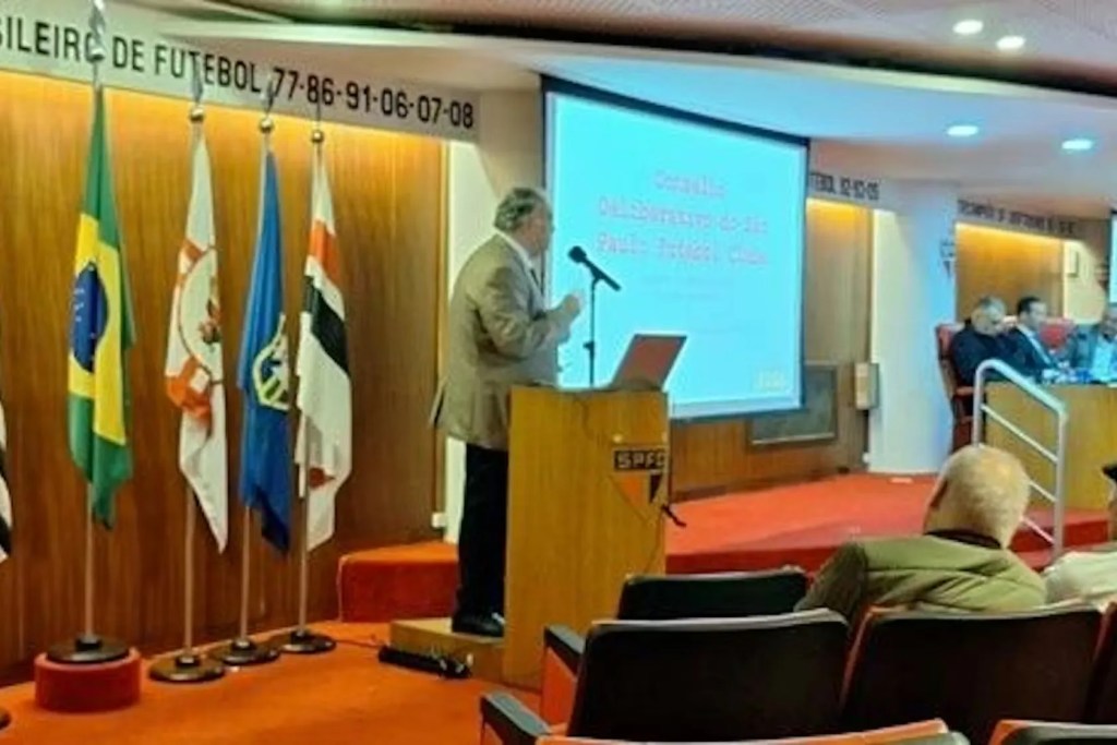 Apresentação em evento de futebol com um palestrante de costas, bandeiras do Brasil e outros estados ao fundo, e uma tela ao lado exibindo informações.