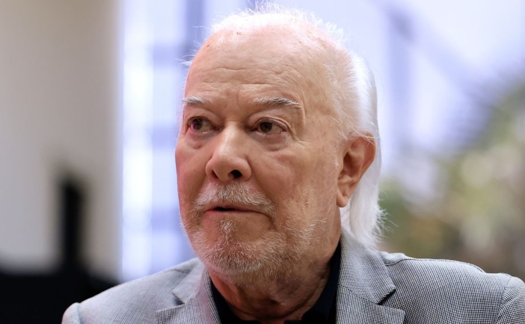Retrato de um homem idoso com cabelo grisalho, usando um blazer claro, em um ambiente interno.