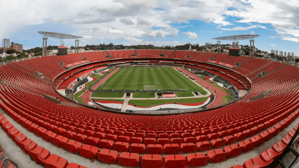 Vista panorâmica do estádio com assentos vermelhos e campo de futebol ao centro, sob um céu parcialmente nublado.