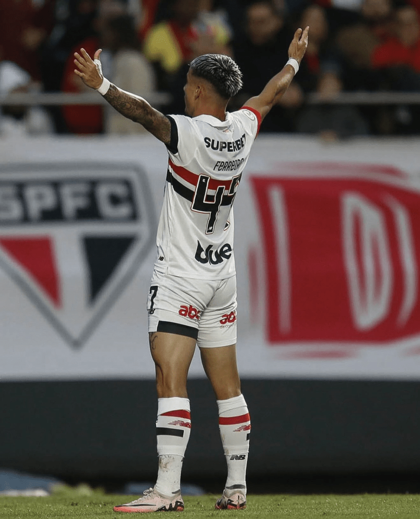 Jogador de futebol do São Paulo FC, usando uniforme branco com detalhes em vermelho e preto, com os braços levantados em sinal de celebração ou convocação durante um jogo.