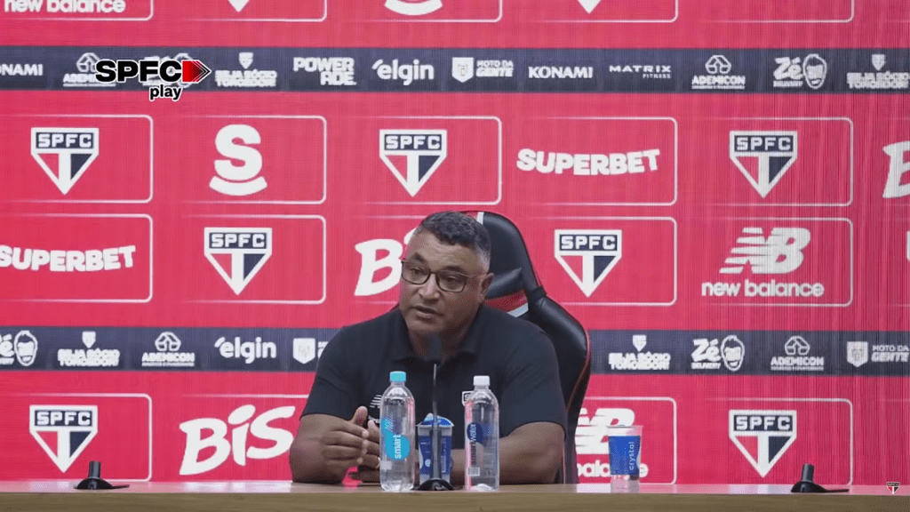 Técnico do São Paulo FC em uma coletiva de imprensa com logotipo do clube ao fundo.