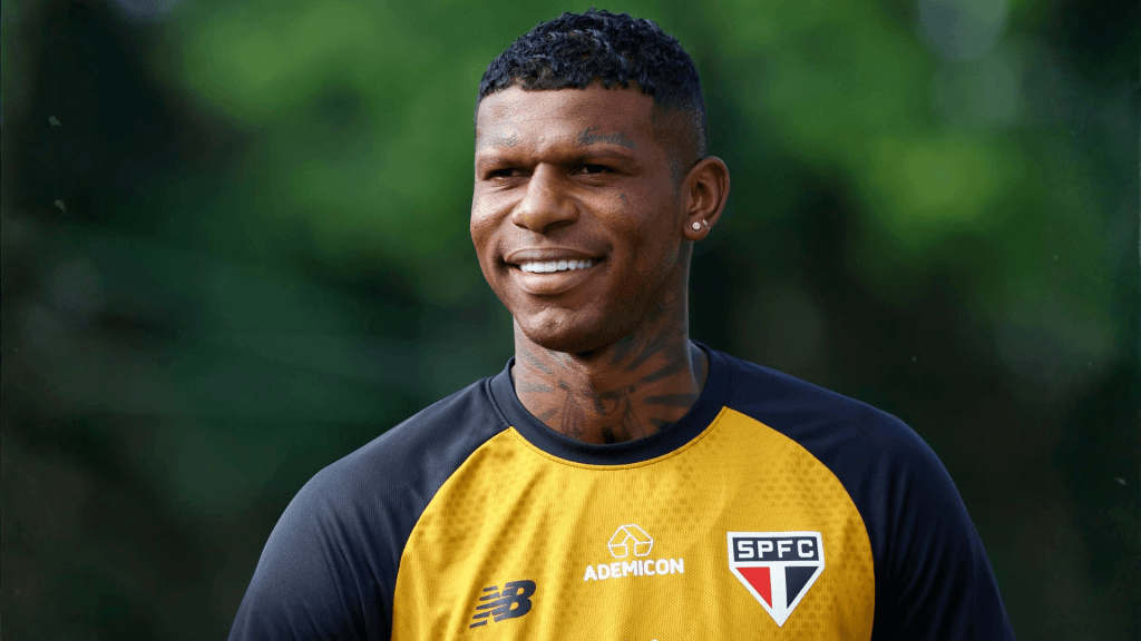 Jogador de futebol sorrindo durante treinamento, vestindo camiseta com logotipo do São Paulo FC.