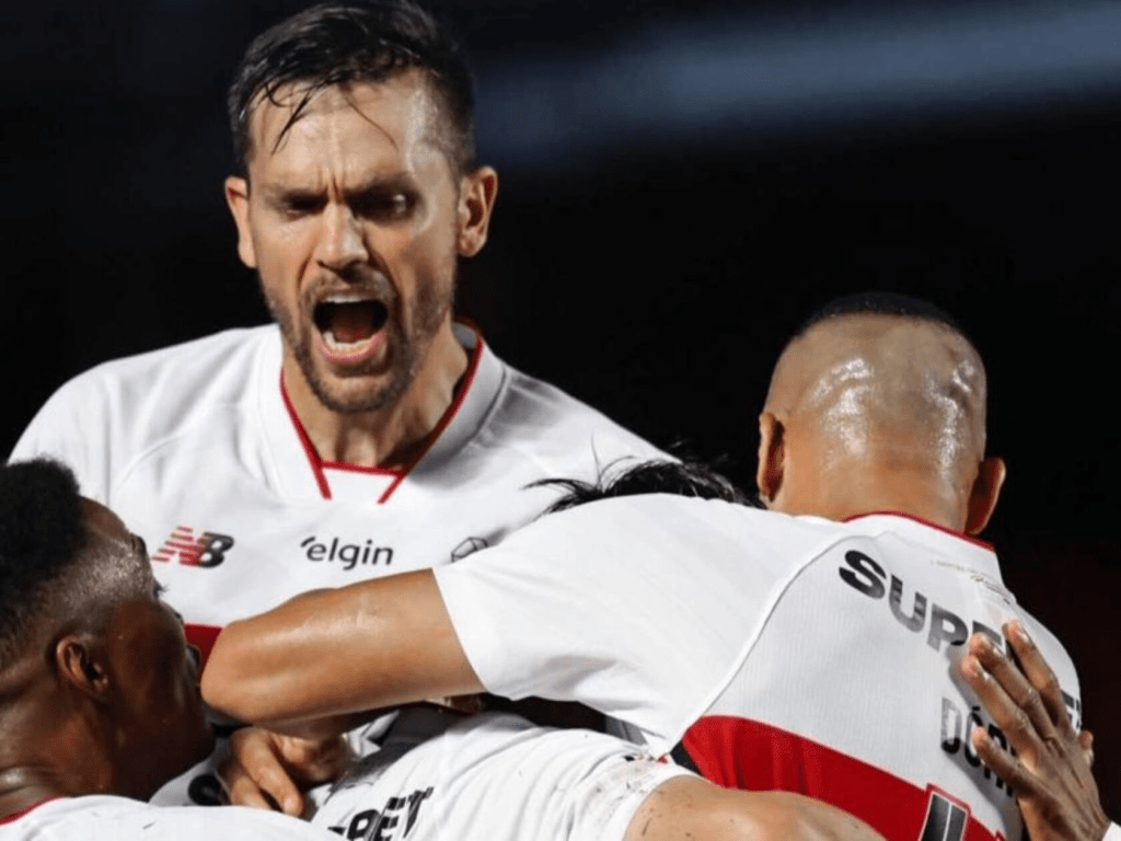 Jogadores de futebol comemorando com entusiasmo após um gol, com um jogador em destaque gritando de alegria.