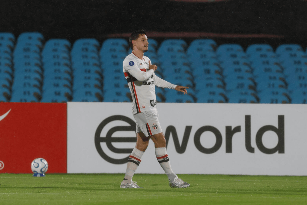 6 Copa Sul Americana 2026 São Paulo FC 1 Jogador de futebol em ação durante uma partida, usando um uniforme branco com detalhes vermelhos e pretos. Ele está apontando para a esquerda, com um fundo de arquibancada azul e um banner de patrocínio ao fundo.