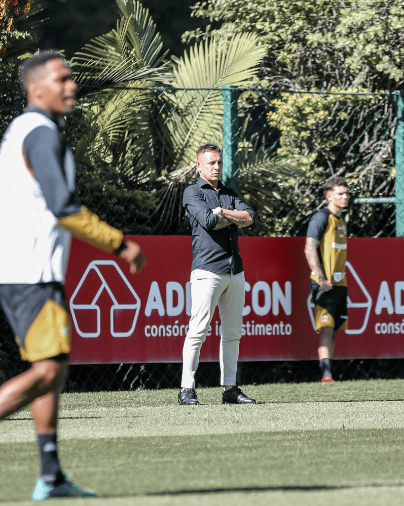 Um treinador em pé em um campo de treinamento, observando jogadores durante a prática. O fundo apresenta um painel publicitário e vegetação ao redor.