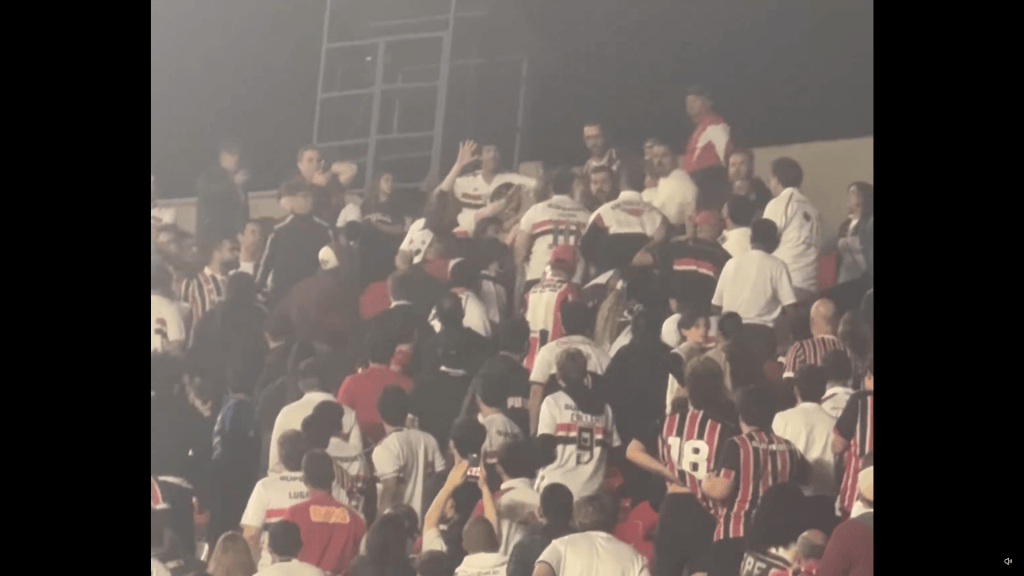 Torcedores em um estádio, muitos vestidos com camisas de futebol, interagindo de forma animada.