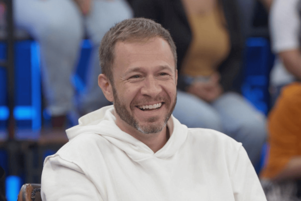 Homem sorrindo, usando um moletom branco, em um ambiente com público ao fundo.
