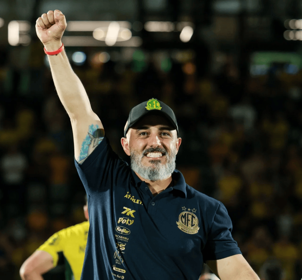 Rafael Guanaes seria uma boa no São Paulo? Perfil do técnico do Mirassol e o que ele poderia oferecer em 2027 1 Homem com barba e cabelo grisalho levantando o punho em sinal de celebração, usando uma camisa azul escura e um boné preto, sorrindo em um ambiente esportivo com torcida ao fundo.